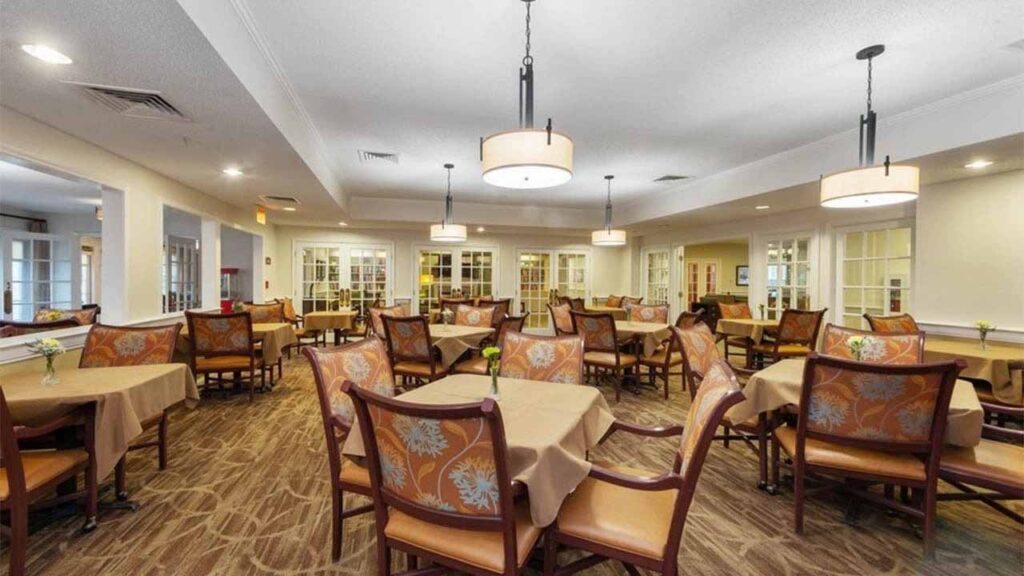 A spacious, well-lit dining room with multiple tables covered in beige cloths, surrounded by wooden chairs with floral-patterned cushions and elegant light fixtures hanging from the ceiling.
