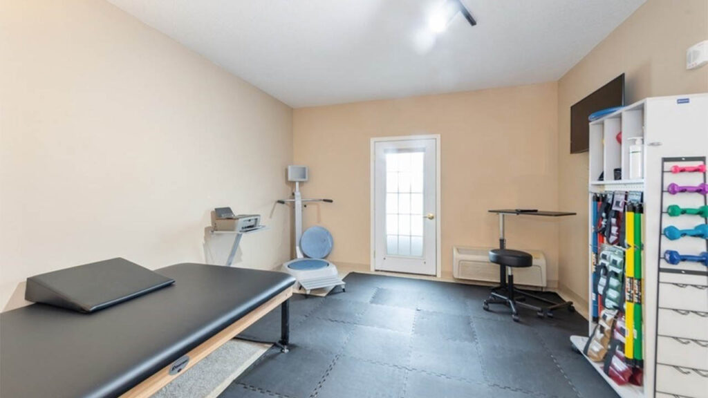 A clean physical therapy room with padded flooring, an exam table, exercise equipment, a standing desk, a wall-mounted TV, and shelves with colorful dumbbells, all against beige walls and a door with a window.