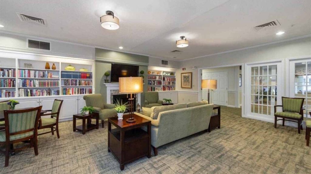 A cozy, well-lit lounge with green armchairs, sofas, wooden tables, lamps, and bookshelves lining the back wall. The carpeted room has light walls, large windows, and a welcoming atmosphere.