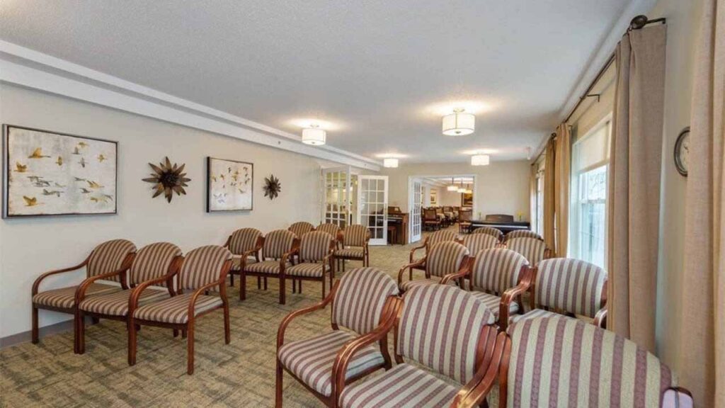 A well-lit room with rows of striped cushioned chairs arranged for an audience, beige carpet, wall art, large windows with curtains, and glass doors at the back.