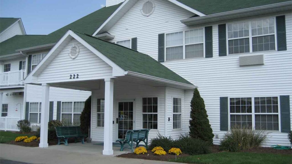 A white, two-story building with green roof and shutters, featuring a covered entrance, benches, and neatly landscaped bushes and yellow flowers in front. The number 222 is displayed above the entryway.