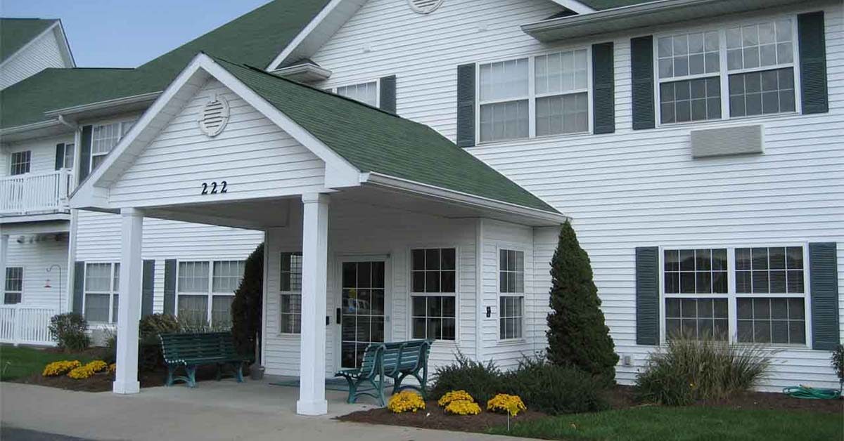 A white two-story building with green roof and shutters, featuring a covered entrance labeled 