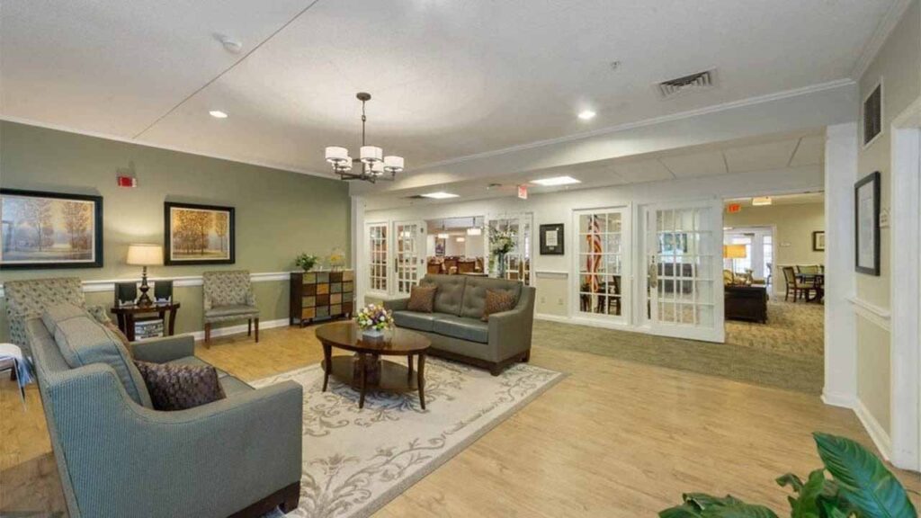 A spacious, well-lit lounge with sofas, armchairs, a coffee table, and decorative plants. Artworks decorate green and white walls, and glass doors open to another cozy room in the background.