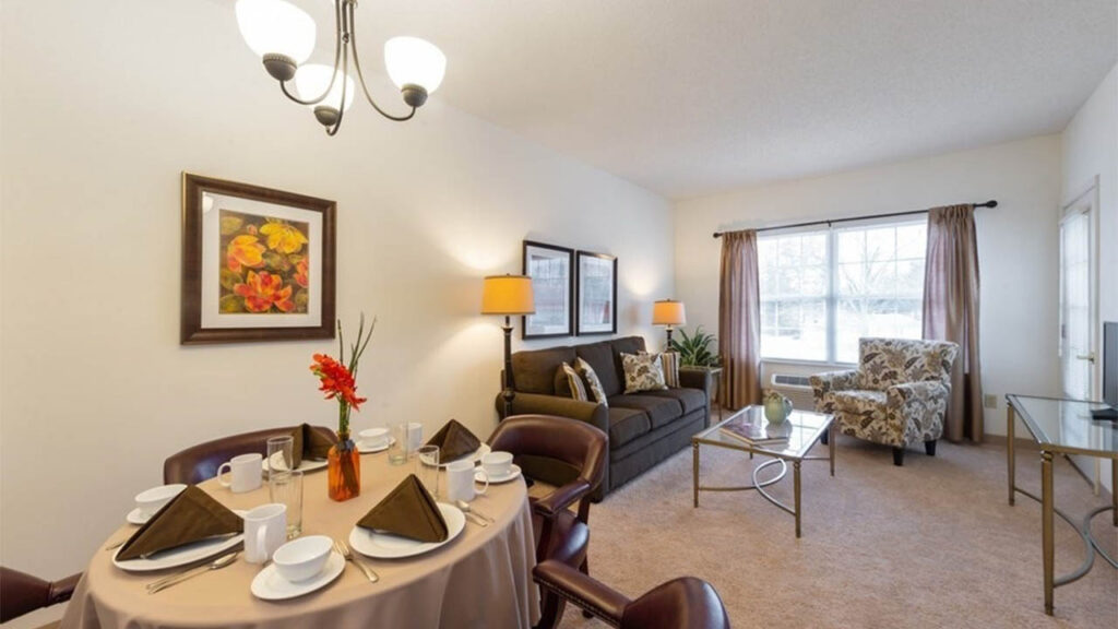 A cozy living and dining room with a round table set for four, a brown sofa, floral armchair, coffee table, wall art, lamps, large window with curtains, and soft beige carpet.