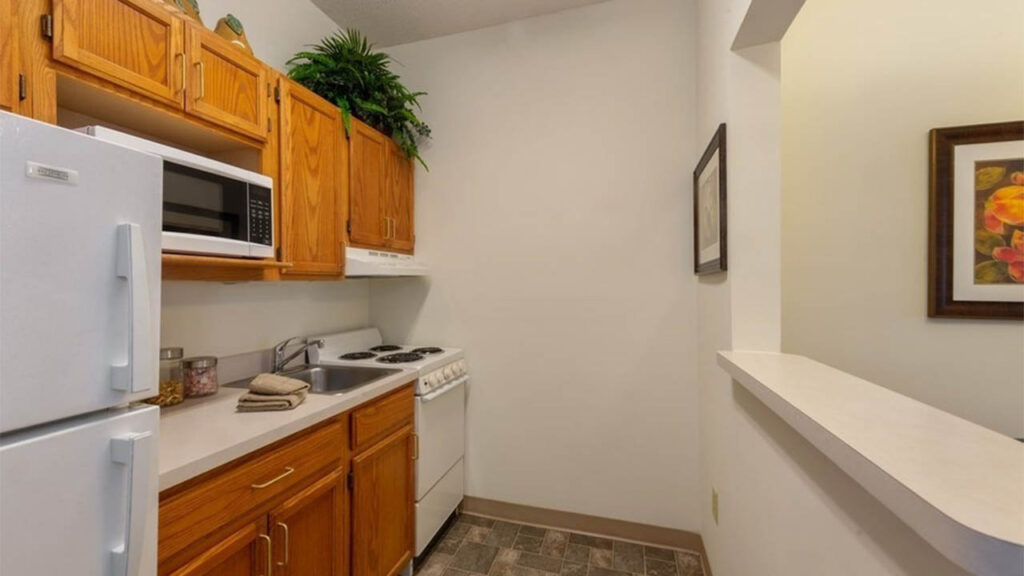 Small kitchen with wooden cabinets, white refrigerator, microwave, stove, and sink. A plant sits above the cabinets. There are framed pictures on the beige walls and a counter dividing the kitchen from another room.