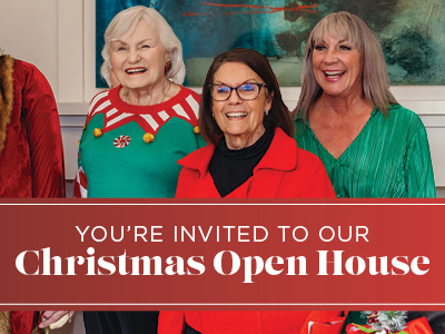 Three smiling older women in festive clothing stand together behind a red banner that reads, 