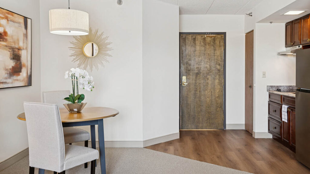 A modern hotel room entryway with a small dining table, two upholstered chairs, a white orchid centerpiece, wall art, a sunburst mirror, and a kitchenette with wooden cabinets and a refrigerator.