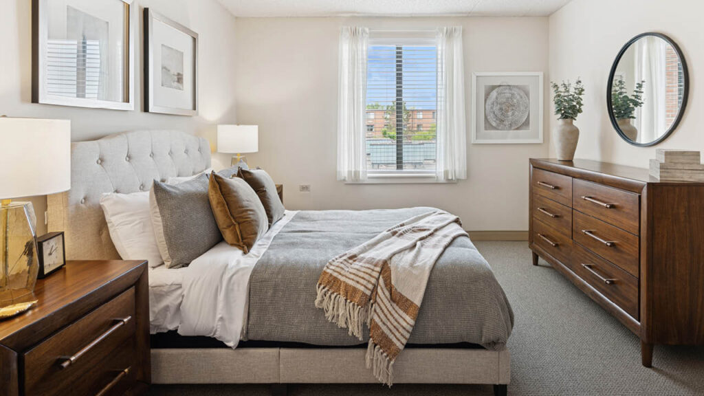 A modern, cozy bedroom with a neatly made bed, earth-toned bedding, wooden nightstand and dresser, round mirror, framed art, potted plants, and a window letting in natural light.