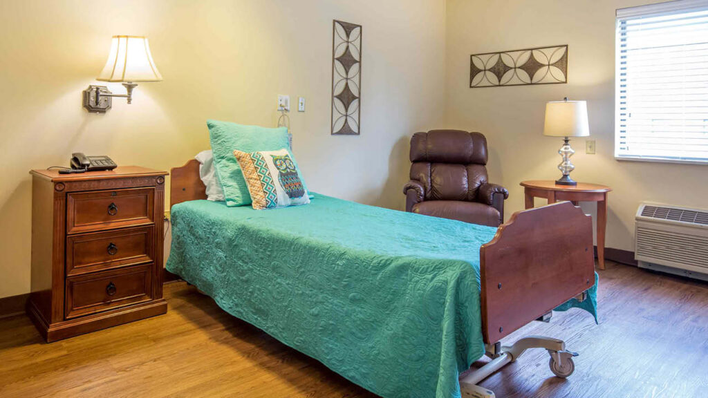 A neatly made single bed with a turquoise cover sits beside a wooden nightstand, brown recliner, and bedside lamp in a cozy, well-lit room with wooden floors and wall decorations.