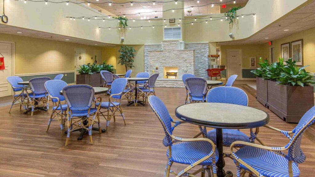 A spacious, well-lit indoor seating area with blue wicker chairs and round tables on wooden flooring, potted plants, string lights overhead, and a stone fireplace on the far wall.