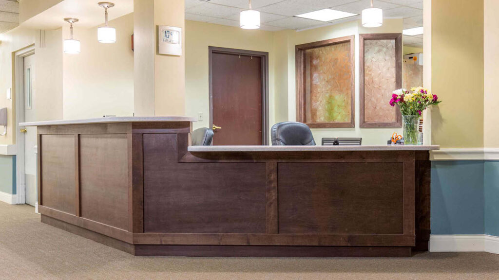 A clean, empty reception desk in an office or clinic lobby with dark wood paneling, two office chairs, a vase of flowers, and abstract art on the walls behind it. Ceiling lights illuminate the area.