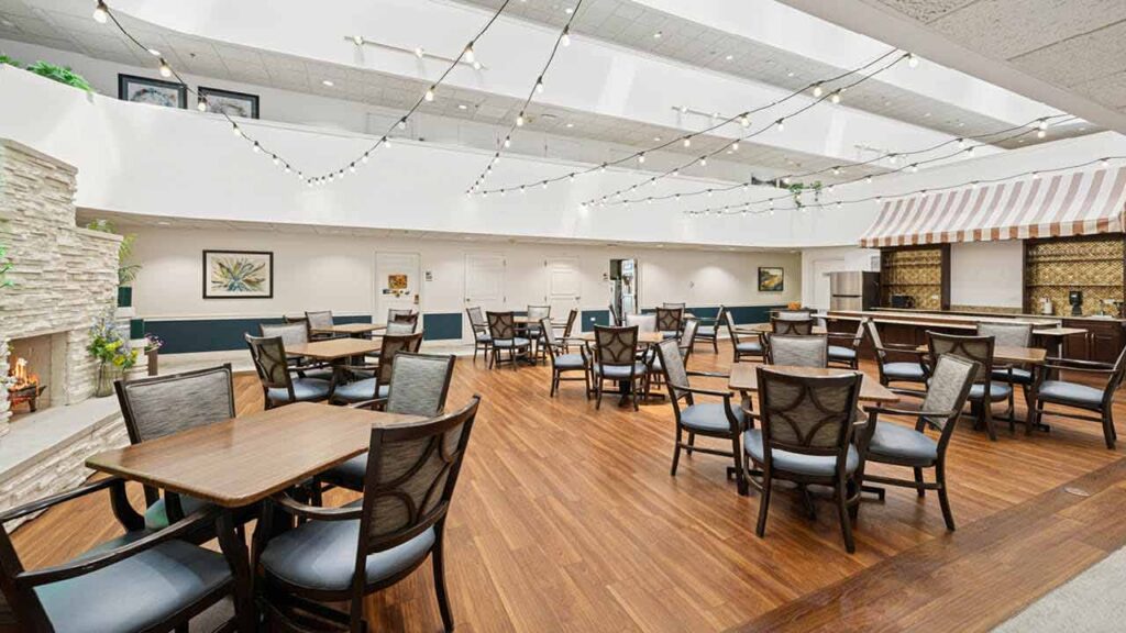 A bright, spacious dining area with wooden floors, several empty tables and chairs, a stone fireplace, string lights overhead, and a counter with a striped canopy on the right side.