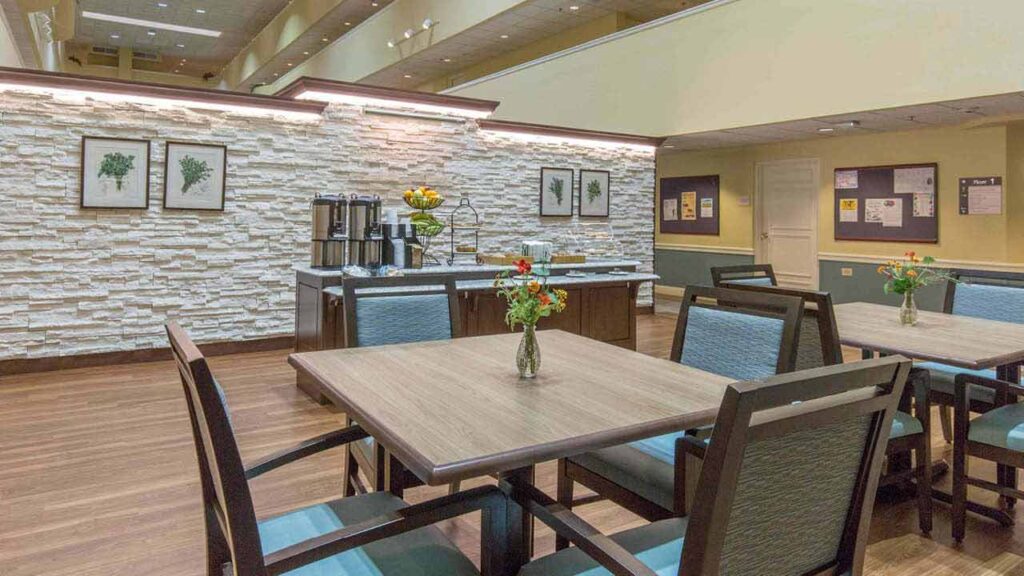 A modern dining area with wooden tables and chairs, flowers in vases on each table, a beverage station against a white brick accent wall, and framed botanical prints on the wall.