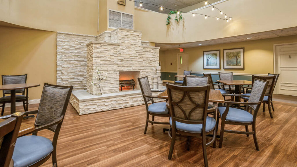 A spacious, well-lit lounge area with wooden floors, several tables and chairs, a large white stone fireplace, framed art on the walls, and string lights hanging from the ceiling.