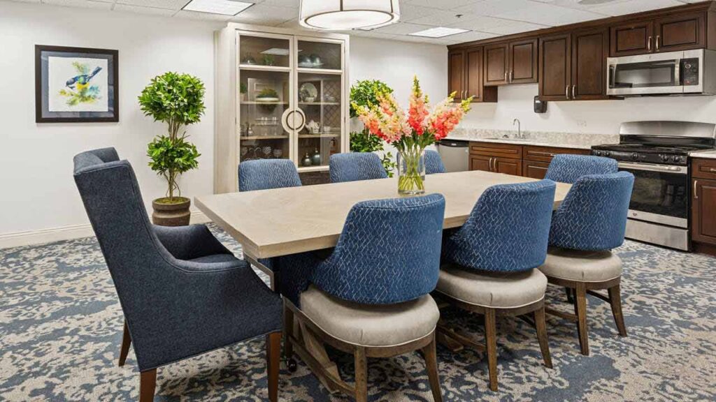 A modern dining room with a large table surrounded by eight blue upholstered chairs, a vase with colorful flowers in the center, two potted plants, a glass-front cabinet, dark wood kitchen cabinets, and stainless steel appliances.