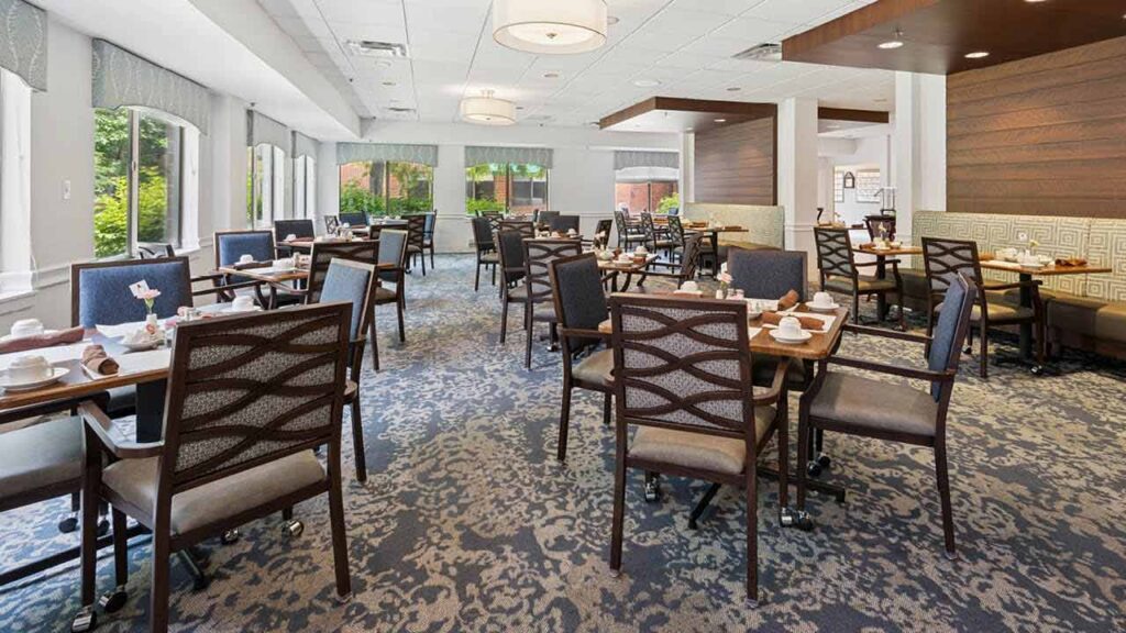 Bright, modern dining room with multiple tables and chairs, set with cups and napkins. Large windows let in natural light, and the room features patterned carpet and neutral-toned decor.