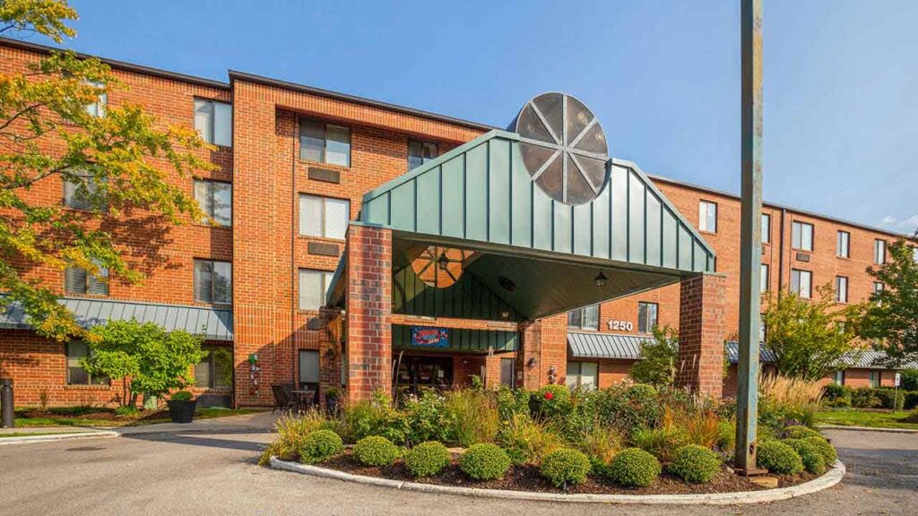 A brick, four-story hotel with a green metal entrance canopy, landscaped garden beds, and large windows under a clear blue sky.
