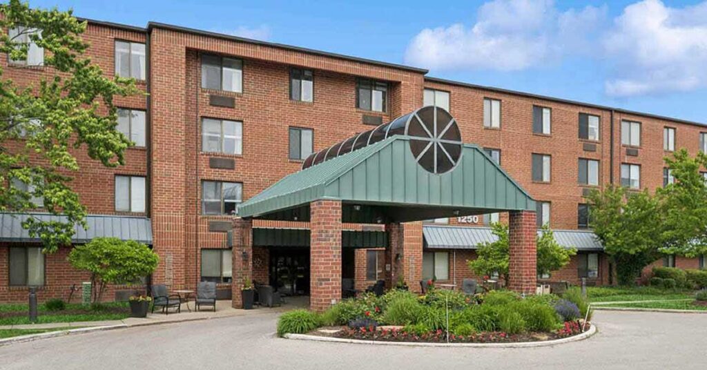 A four-story brick apartment building with a green metal entrance canopy, surrounded by landscaped plants and trees, under a partly cloudy blue sky.
