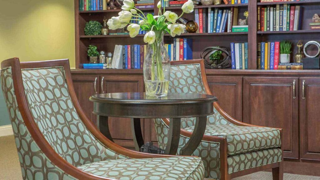 Two patterned armchairs and a round wooden table with a vase of white tulips are in front of a wooden bookcase filled with colorful books and decorative items.