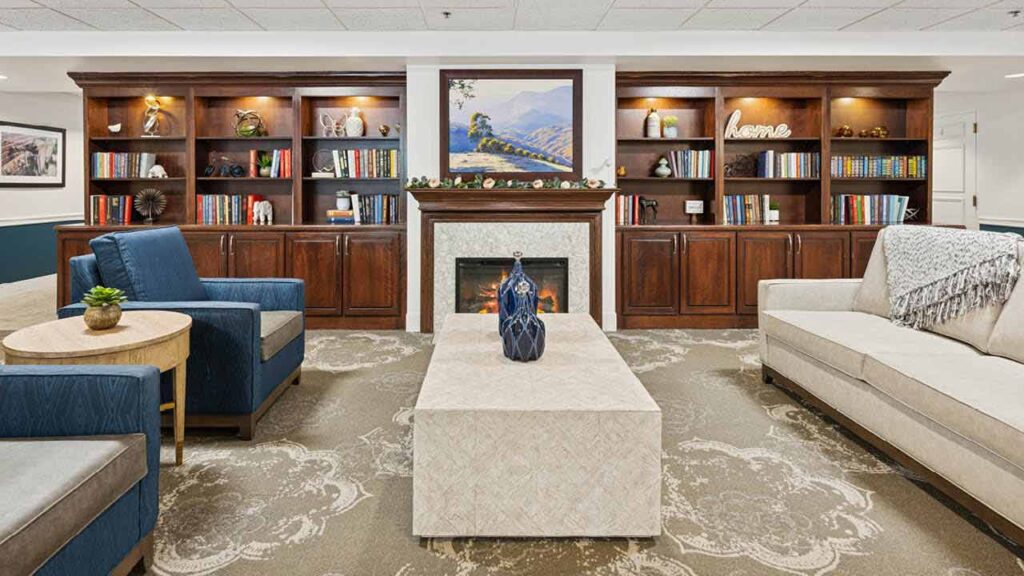 A cozy lounge with a beige sofa, blue armchairs, a light patterned rug, and a marble coffee table. Behind, bookshelves and a fireplace are topped with a landscape painting and decorative items.