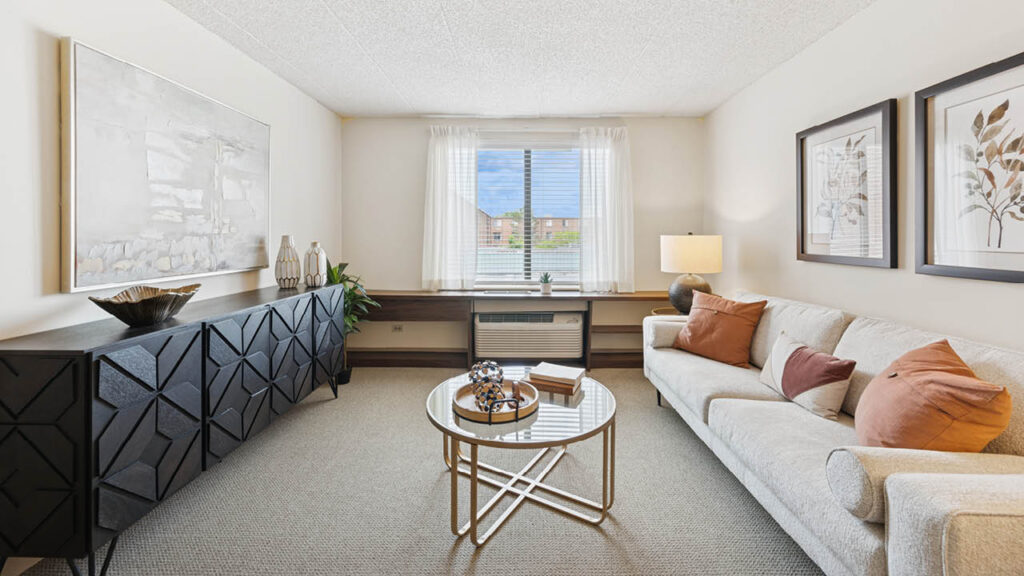 A bright living room with a beige sofa, decorative pillows, a round glass coffee table, modern art on the walls, a black sideboard with vases, and a large window with sheer curtains letting in natural light.