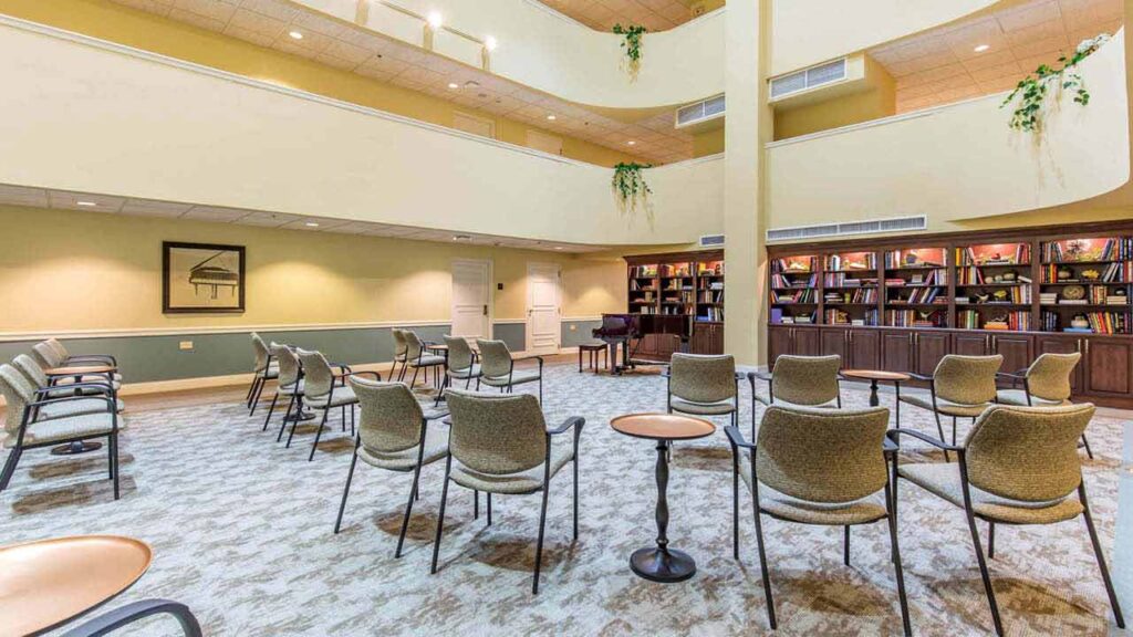 A spacious, well-lit room with rows of empty chairs arranged in a semicircle, small round tables, a grand piano, bookshelves filled with books, and green plants on upper ledges.