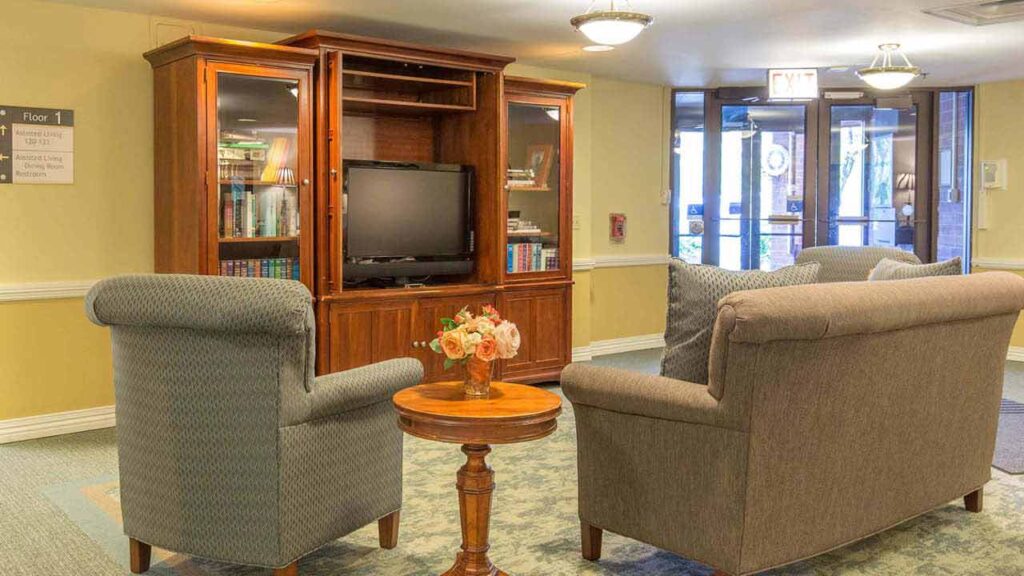 A cozy lounge area with two upholstered chairs facing a wooden TV cabinet, bookshelves, a small round table with a flower arrangement, and large glass doors in the background letting in natural light.