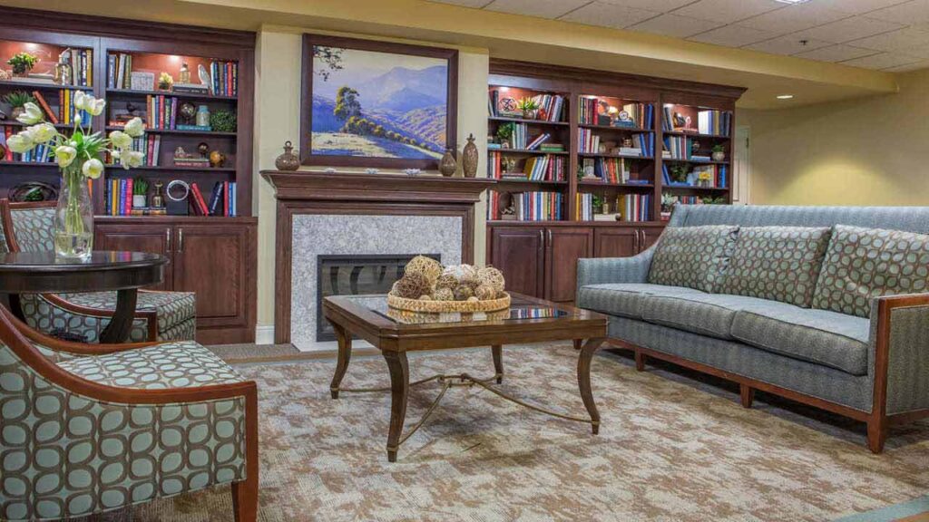 A cozy living room with a sofa, armchair, coffee table, fireplace, bookshelves filled with books, a flower vase, and a landscape painting above the mantel.