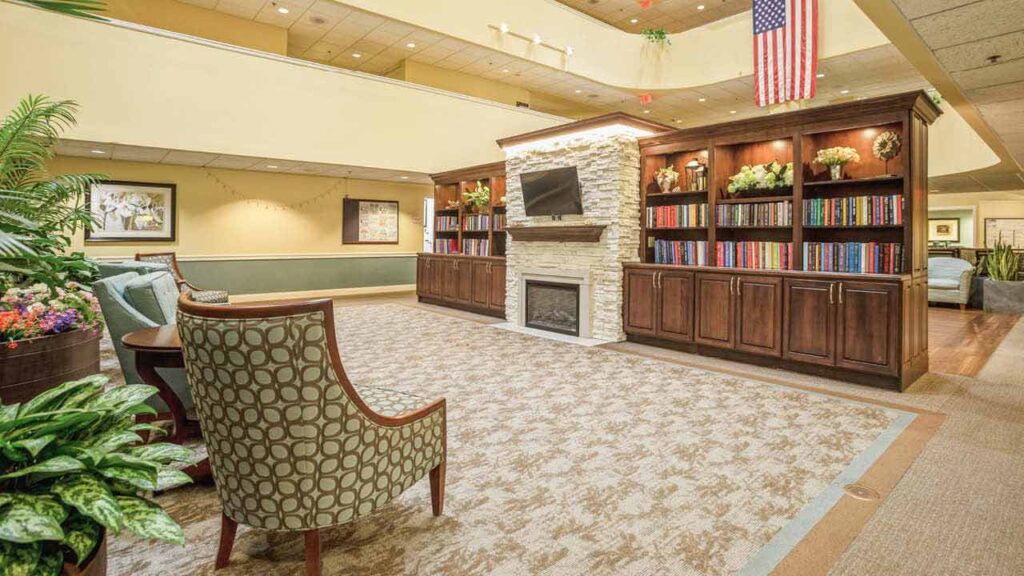 A spacious, well-lit lobby with a patterned carpet, cushioned chairs, plants, built-in bookshelves, a fireplace, a TV, and an American flag hanging from the ceiling.