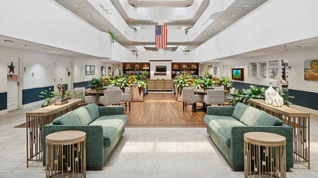 Spacious, well-lit lobby with two green sofas, wooden tables, many plants, and an American flag hanging above. A seating area and a bar or reception desk are visible in the background.