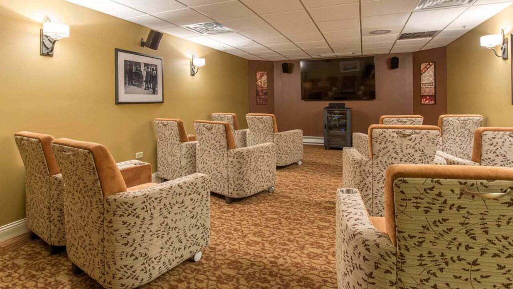 A small home theater room with two rows of beige patterned armchairs facing a large wall-mounted TV, soft wall lighting, framed art, and a carpeted floor.