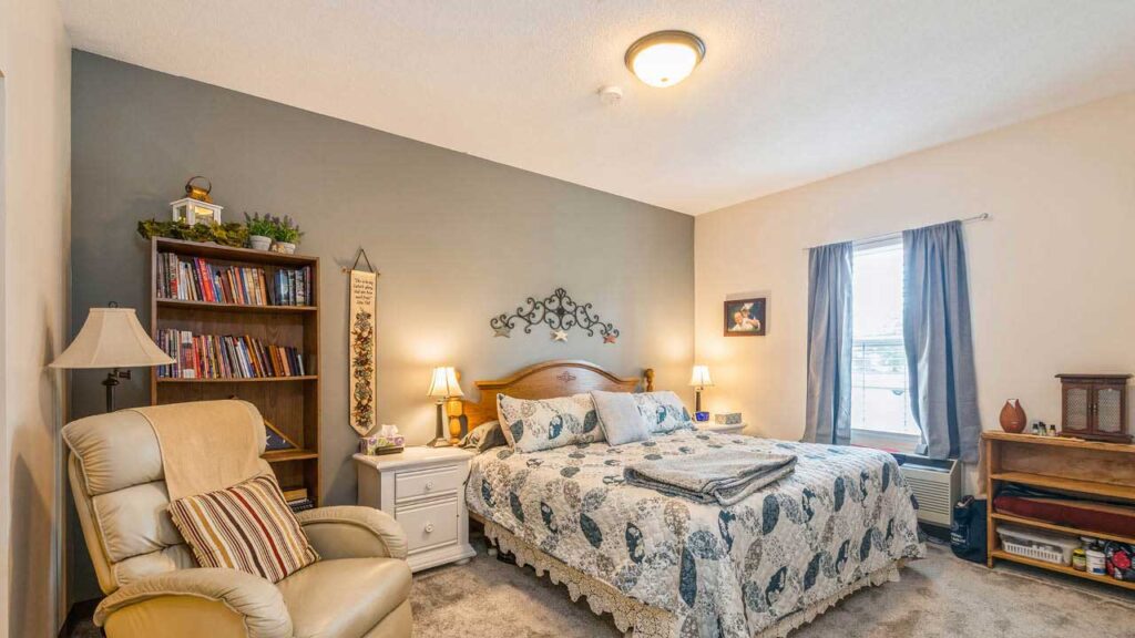 A cozy bedroom with a bed covered in a patterned quilt, two bedside tables with lamps, a bookshelf filled with books, an armchair, and a window with gray curtains letting in natural light.
