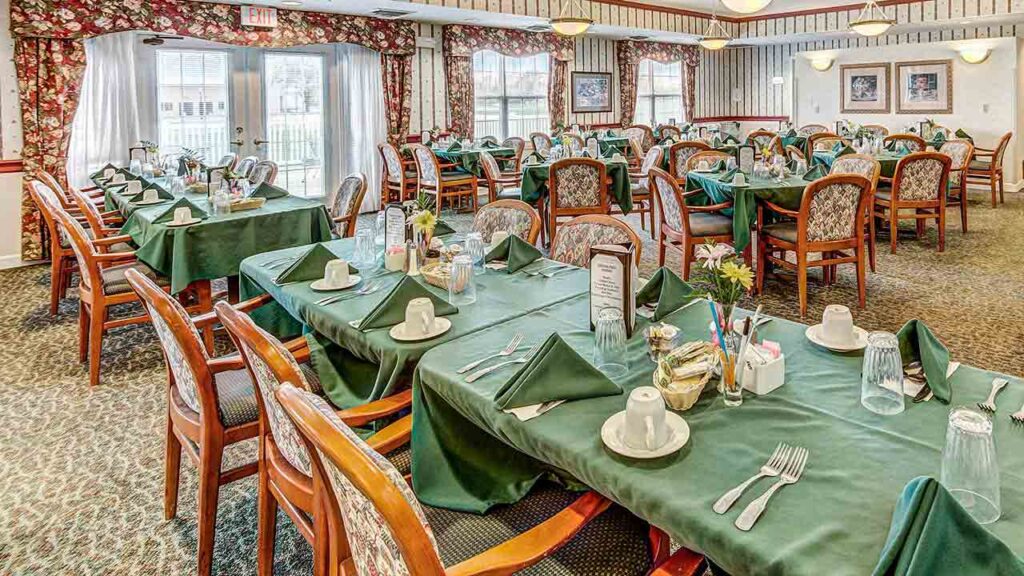 A spacious dining room with multiple tables set for a meal, featuring green tablecloths, wooden chairs, floral curtains, and large windows letting in natural light. Each table is neatly arranged with tableware and napkins.