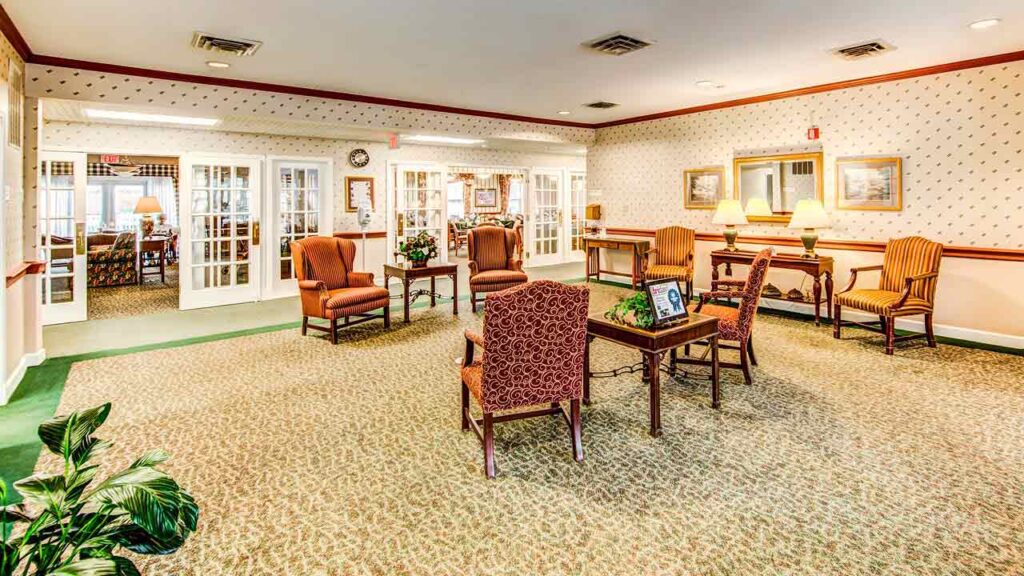 A cozy, well-lit lounge with patterned carpet, upholstered chairs, side tables with lamps, framed art on wallpapered walls, and glass doors leading to another room. Houseplants decorate the space.