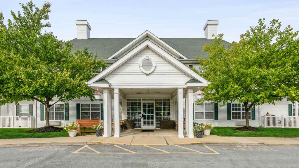 A front view of a white, single-story building with a green roof, large covered entrance, double glass doors, and trees on either side. Benches and potted plants decorate the entryway.
