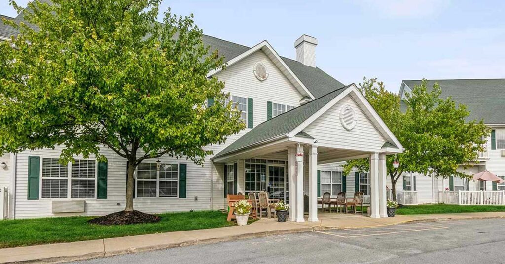 A two-story white building with green shutters and a covered entrance, surrounded by trees and outdoor seating, resembles an assisted living or senior care facility. The sky is clear and the grounds are well maintained.