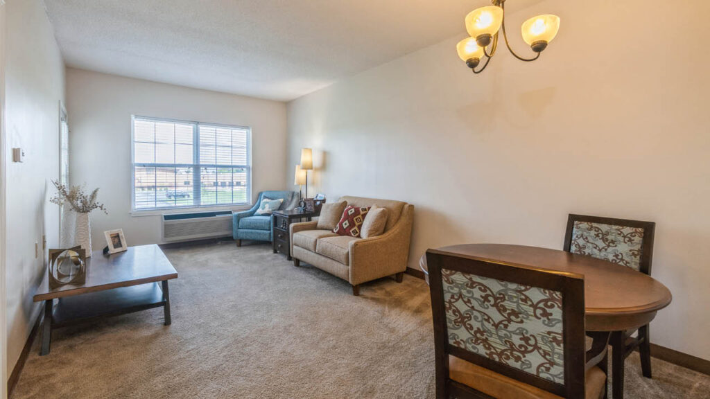A cozy living room with beige carpet, a small dining table with two chairs, a loveseat, an armchair, a coffee table, and a window letting in natural light. A wall-mounted light fixture hangs above the table.