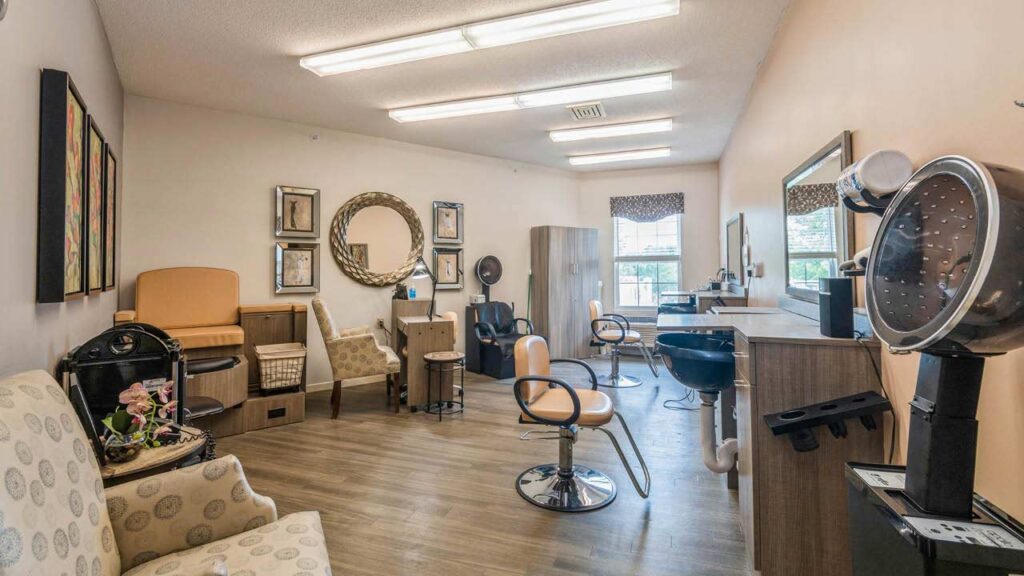 A well-lit hair salon with multiple chairs, mirrors, and styling stations. The space features framed artwork, a patterned armchair, hair dryers, and large windows letting in natural light.
