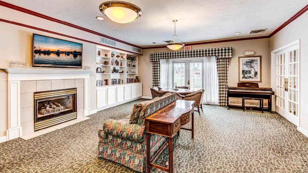 A cozy living room with a fireplace, floral sofa, wooden table, wall shelves with books, a TV above the mantel, a piano in the corner, and glass doors leading outside. The room has patterned carpet and elegant lighting.