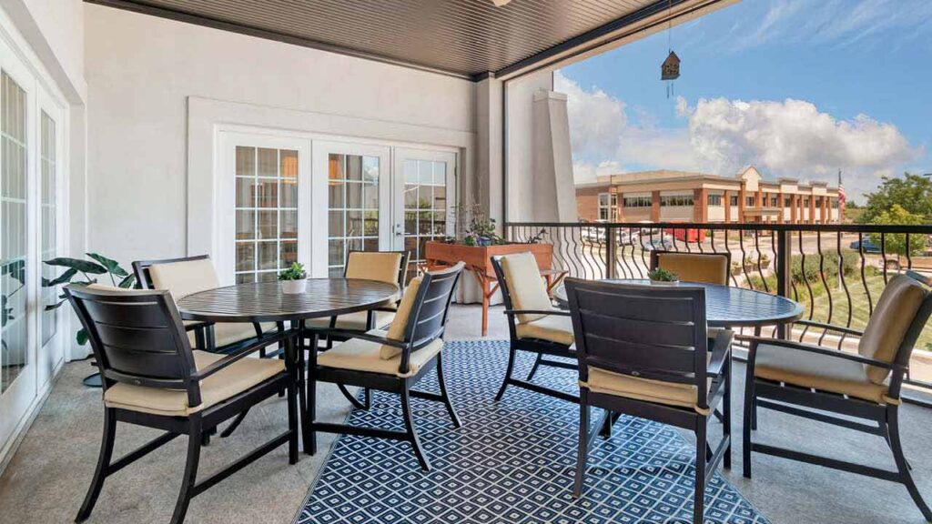 Spacious covered patio with two round tables, several chairs, potted plants, a blue patterned rug, and a view of a building and sky in the background.