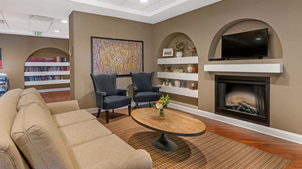 A cozy lounge with a tan sofa, two dark armchairs, a round wooden coffee table, a fireplace, wall shelves with decor, a flat-screen TV, and colorful artwork. A bookshelf is visible in the background.