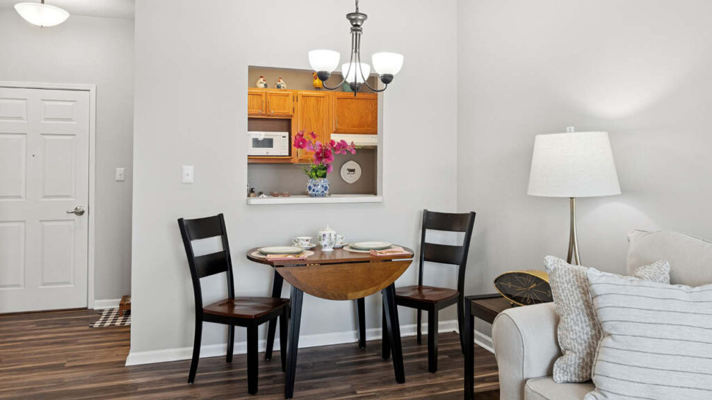 A small dining area with a wooden table set for two, two black chairs, a floor lamp, and a view through a wall opening into a kitchen with wooden cabinets and a vase of pink flowers.