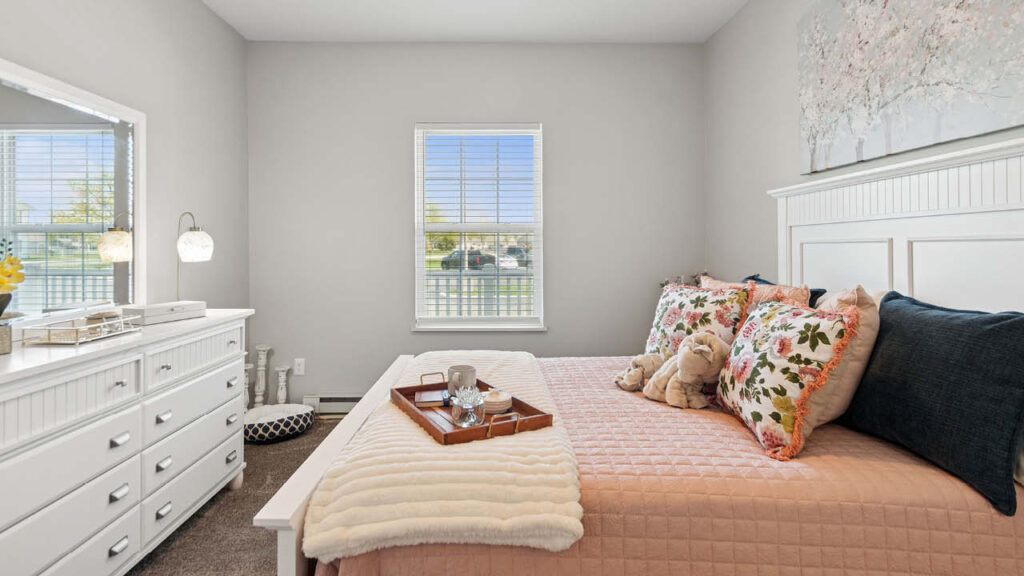 A cozy bedroom with a white dresser and mirror, a bed with pink bedding, floral pillows, stuffed animals, and a tray. A window lets in natural light, and the room features soft, neutral decor.