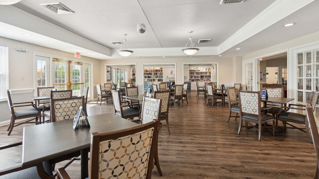 A spacious, well-lit room with several tables and chairs, wooden flooring, and large windows. Bookshelves line the back wall, and modern light fixtures hang from the ceiling, creating a welcoming, communal atmosphere.