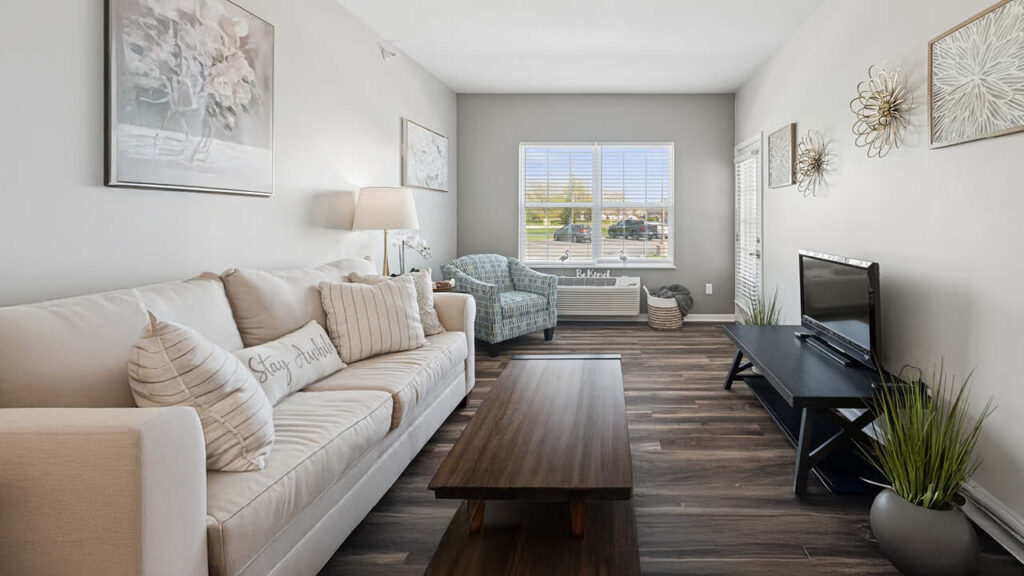 A bright living room with a beige sofa, patterned armchair, wooden coffee table, TV on a black stand, wall art, plants, and a large window letting in natural light. The decor is modern and cozy.