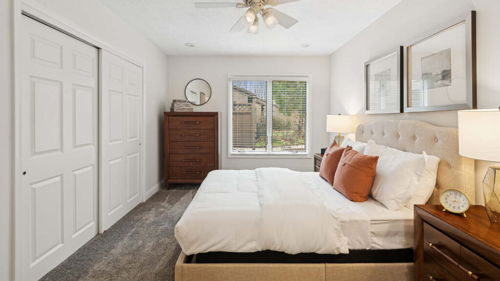 A neatly made bed with white bedding and brown accent pillows sits in a bright bedroom with beige walls, wooden furniture, two bedside lamps, and a window letting in natural light.