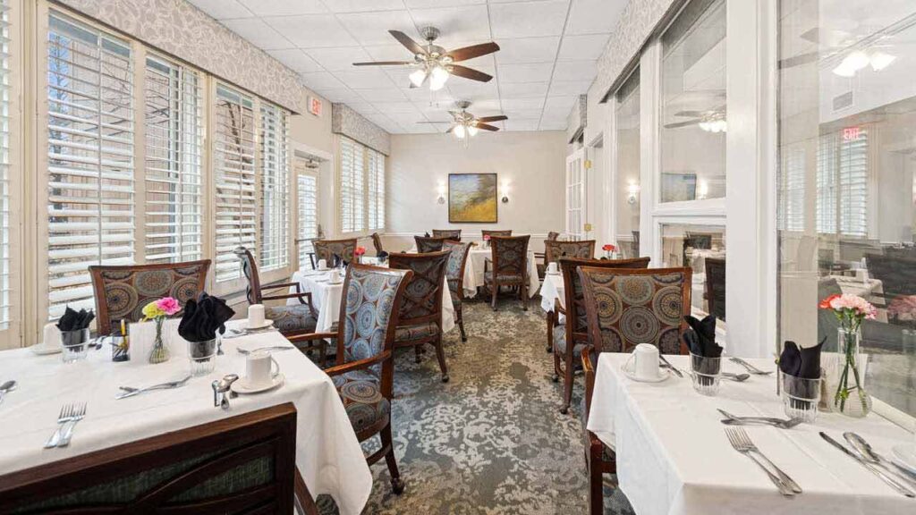 A bright, elegant dining room with several tables covered in white cloths, set with cups, cutlery, and flower vases. Patterned chairs surround the tables. Ceiling fans and large windows let in natural light.