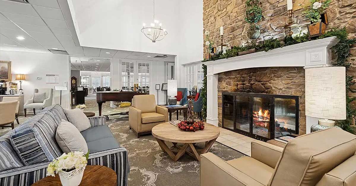 A cozy living room with a lit stone fireplace, comfortable sofas and armchairs, a round wooden coffee table with decor, and greenery on the mantle; a well-lit, inviting space with neutral and warm tones.
