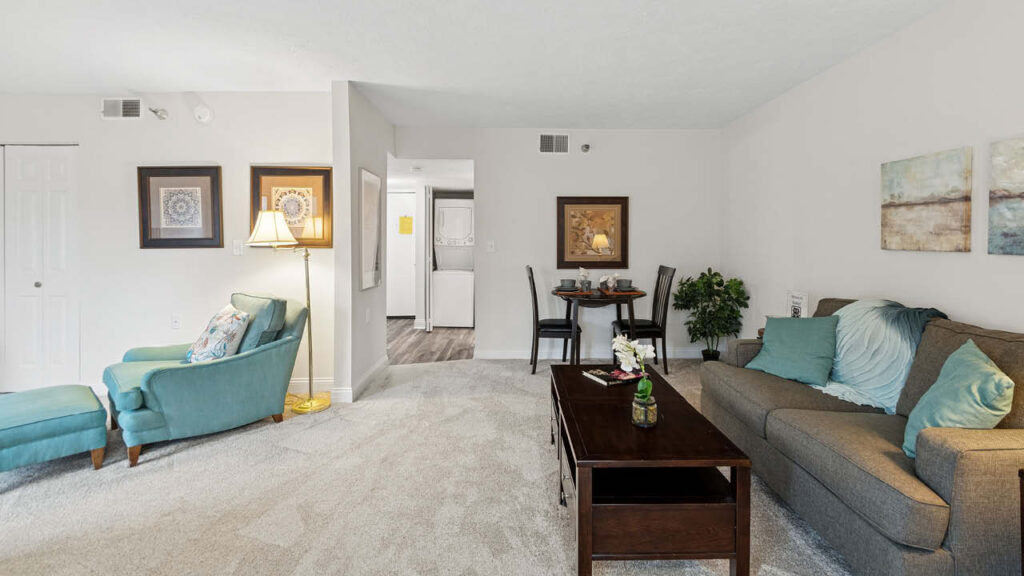 A cozy living room with a teal armchair, a brown sofa with light blue pillows, a wooden coffee table, framed art on the walls, a floor lamp, and a small dining area with two chairs in the background.