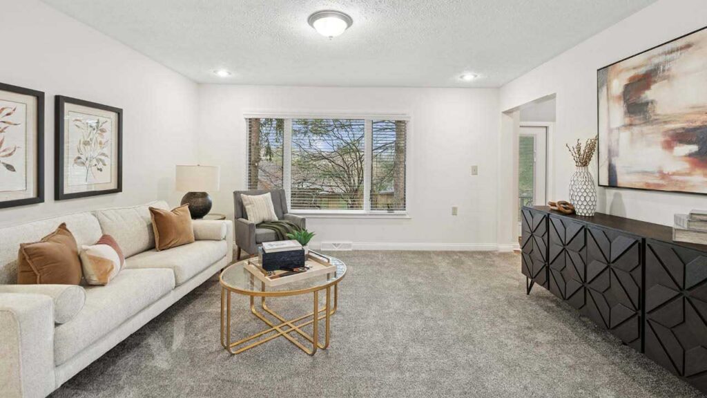A modern living room with a light sofa, accent pillows, a gray armchair, a round glass coffee table, and a black geometric sideboard. Large window with trees outside, and abstract art on the walls.